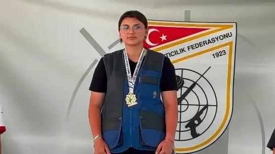 Gözlüklü bir genç, üzerinde madalyalar ve atış yeleğiyle Türkiye Atıcılık Federasyonu logosu önünde duruyor.