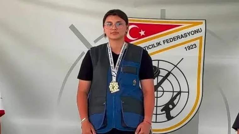 Gözlüklü bir genç, üzerinde madalyalar ve atış yeleğiyle Türkiye Atıcılık Federasyonu logosu önünde duruyor.