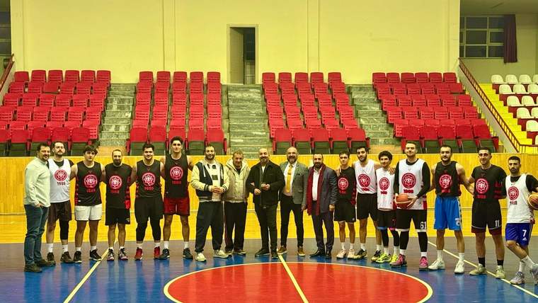 Kapalı spor salonunda basketbol formaları giyen oyuncular ve birkaç kişi, saha ortasında yan yana duruyor.