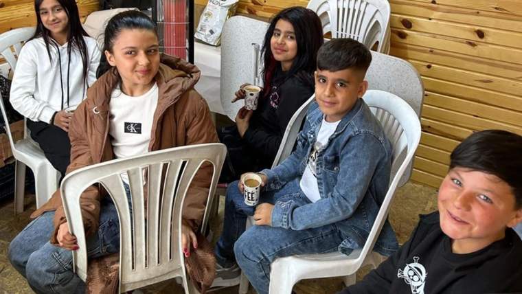 Ahşap duvarlı bir salonda beş çocuk, beyaz plastik sandalyelerde otururken ellerinde küçük bardaklar tutuyor.