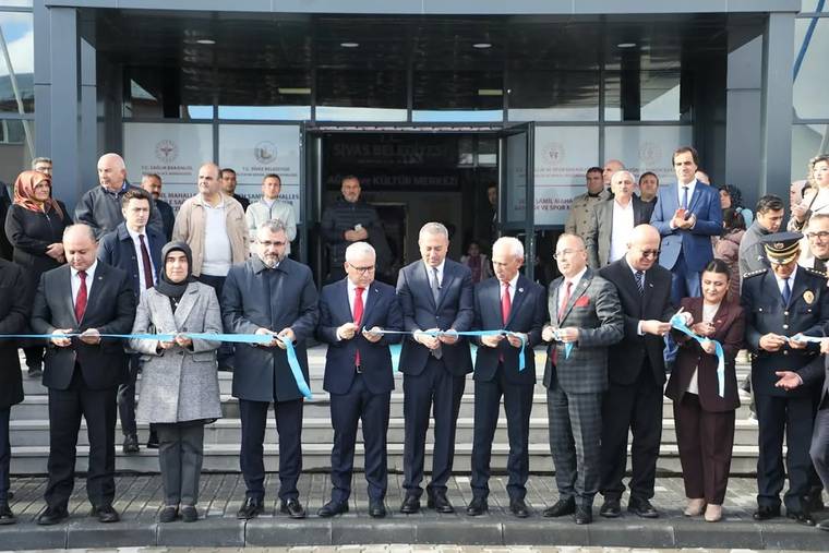 Bina girişinde kalabalık bir grup, mavi kurdeleyi makaslarla keserken yan yana duruyor.