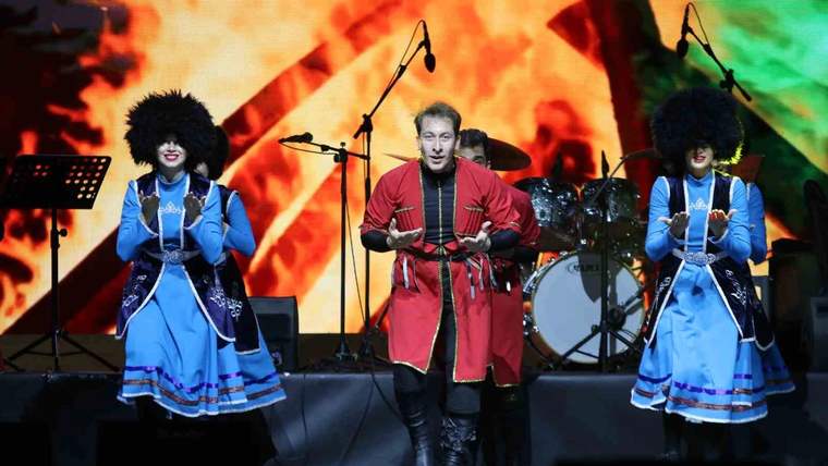 Sahnede geleneksel kıyafetler giyen üç kişi, mikrofonlar ve davul eşliğinde performans sergiliyor.