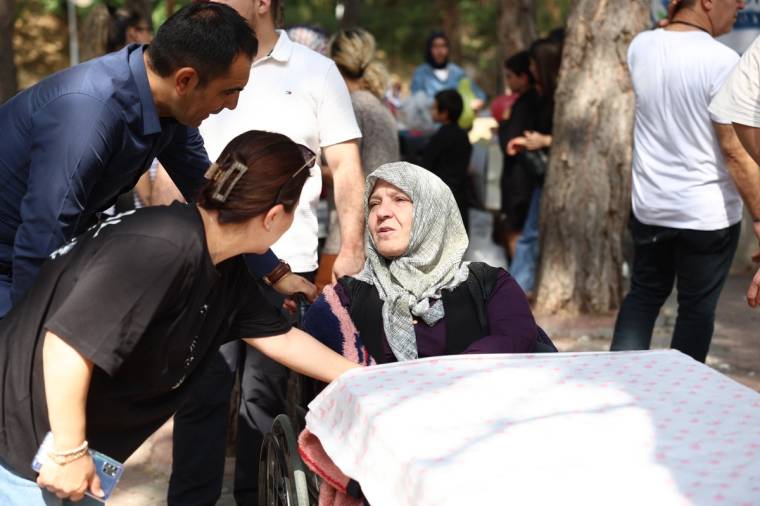 Başörtülü yaşlı bir kadın, açık havada tekerlekli sandalyede otururken iki kişi onunla ilgileniyor.