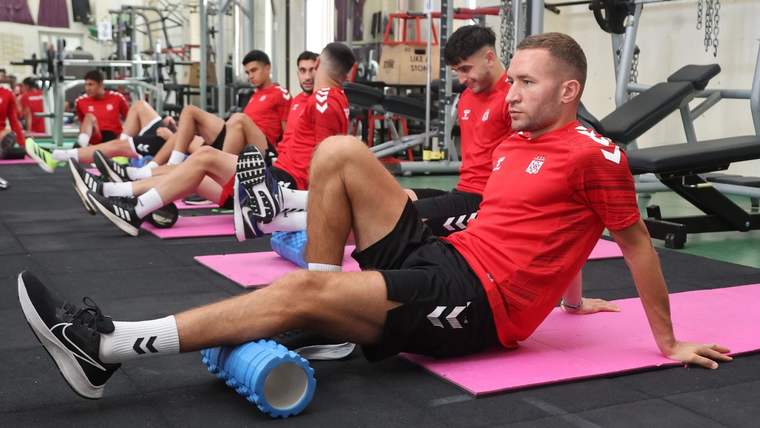 Spor salonunda kırmızı formalı birkaç kişi pembe matlar üzerinde esnerken bir kişi köpük rulosu kullanıyor.