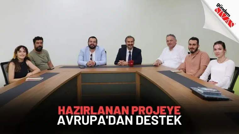 Yedi kişi, toplantı masası çevresinde duvar önünde oturuyor; çoğu kameraya bakıyor.