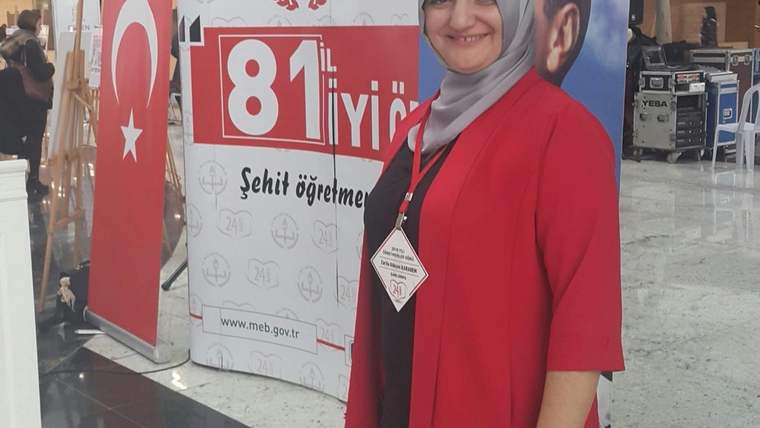 Gri başörtülü ve kırmızı kıyafetli bir kadın, üzerinde “81 il” yazan afişin önünde duruyor.