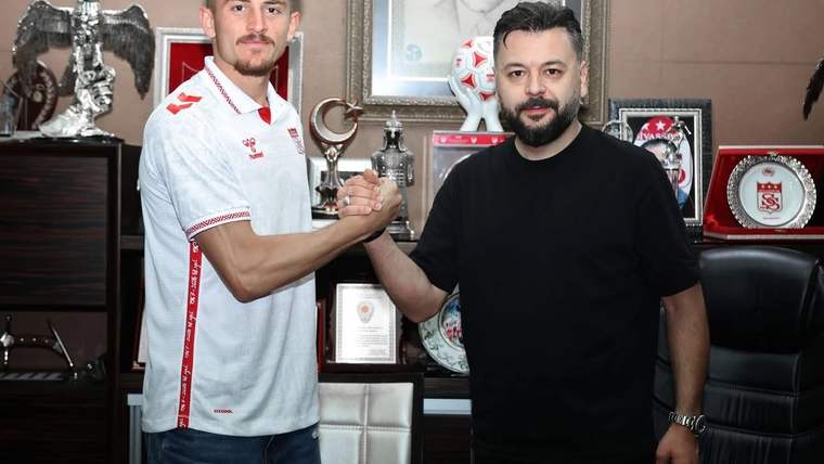 Bir futbolcu, siyah tişörtlü bir kişiyle el sıkışıyor; arka planda kupa ve ödüller var.