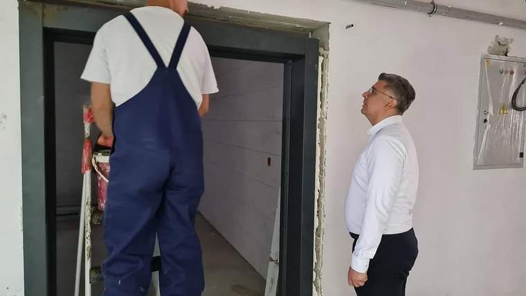 İnşaat halindeki iç mekânda tulumlu bir işçi ve beyaz gömlekli bir kişi, asansör girişinin yanında duruyor.
