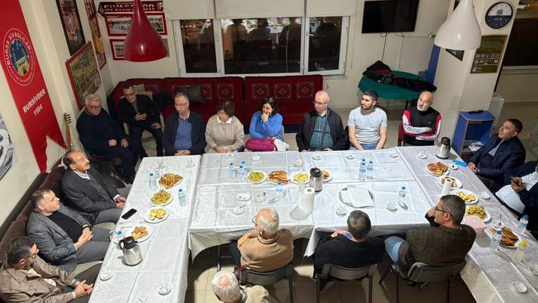 Bir grup insan, yuvarlak bir masa etrafında oturmuş, sohbet ediyor. Masada yiyecek ve içecekler var.