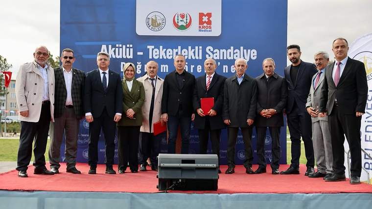 Bir grup insan, açık alanda düzenlenen bir etkinlikte sahnede duruyor. Arkada büyük bir afiş var.