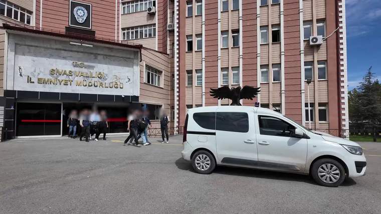 Üzerinde Sivas Valiliği ve İl Emniyet Müdürlüğü yazan bina önünde beyaz bir minibüs ve insanlar görülüyor.
