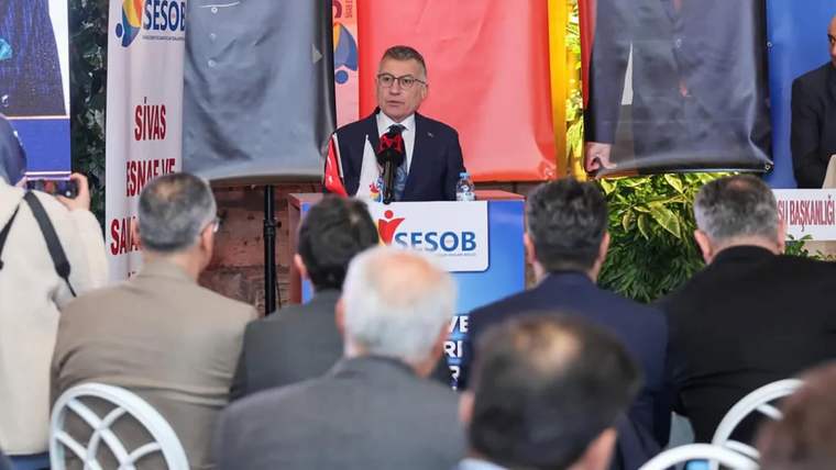 Mikrofon önünde konuşan bir kişi, ESOB yazılı kürsünün arkasında otururken salondakiler onu dinliyor.