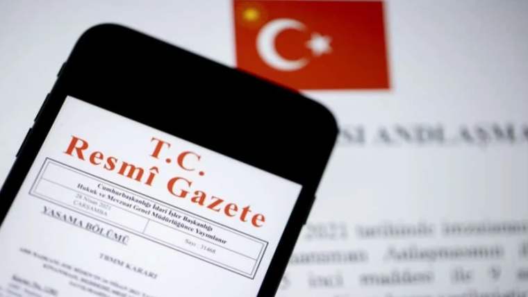 Bir cep telefonunun ekranında T.C. Resmî Gazete belgesi görünüyor, arka planda Türk bayrağı var.