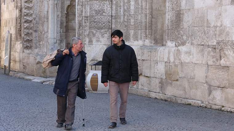 İki kişi, tarihi bir yapının önünde yürüyerek sohbet ediyor.