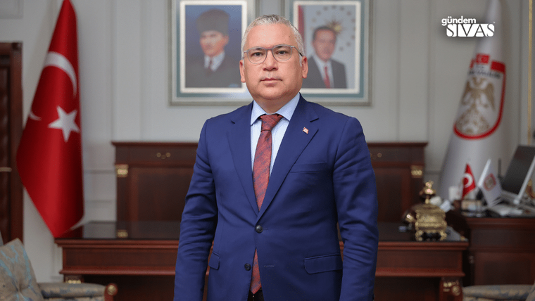 Takım elbiseli bir kişi, Türk bayrağı ve flamaların bulunduğu bir ofiste kameraya bakıyor.