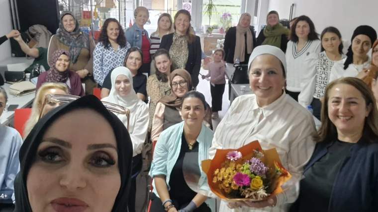 Bir grup kadın, konferans alanında gülümseyerek poz veriyor; ön planda çiçek buketi tutan bir kadın var.