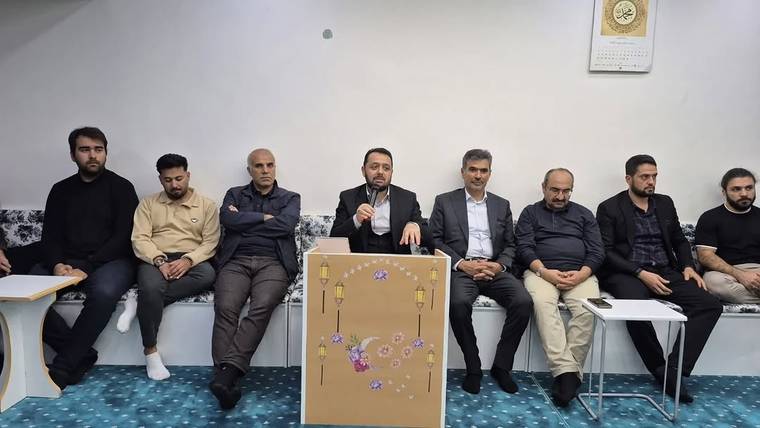 Bir odada yan yana oturan erkekler arasında, ortadaki kişi mikrofonla konuşurken önünde süslü pano duruyor.