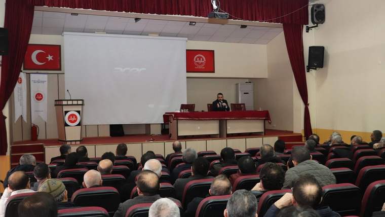 Bir toplantı salonunda, sahnede bir kişi konuşuyor, dinleyiciler sıralarda oturuyor.