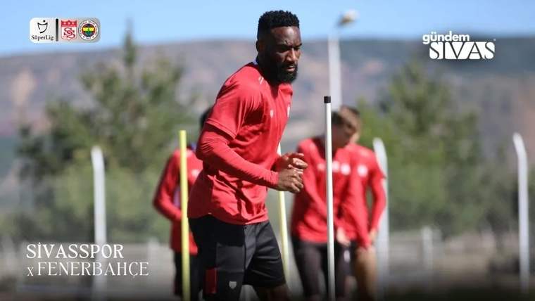 Kırmızı formalı bir futbolcu, antrenman sahasında çubukların arasında koşarken; arka planda iki futbolcu duruyor.