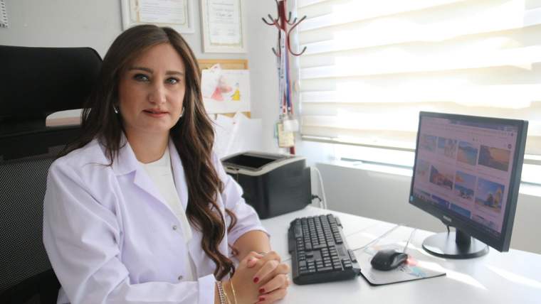 Beyaz önlüklü bir kadın, masa başında bilgisayar ve klavye yanında oturuyor.