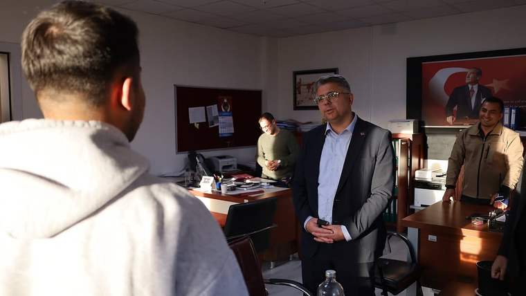 Ofis içinde bir adam, karşısındaki gence bakarken; masaların arasında üç kişi daha ayakta duruyor.
