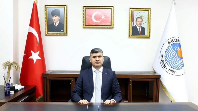 Takım elbiseli bir kişi çalışma masasının arkasında oturuyor; yanında Türk bayrağı ve Akdeniz Belediyesi bayrağı bulunuyor.