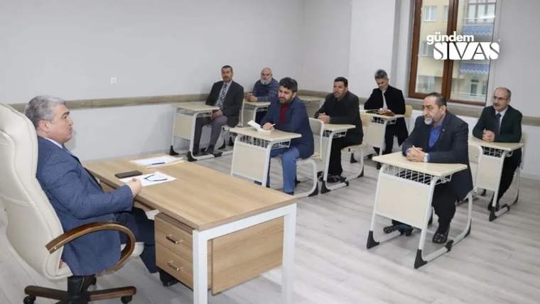 Sınıfta masalarda oturan yetişkinler, önde oturan bir kişinin karşısında ders dinliyor.