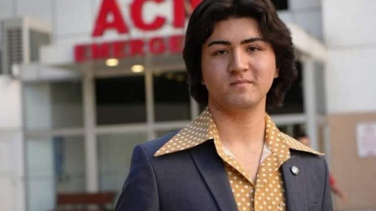 Kahverengi bir ceket ve sarı benekli bir gömlek giymiş genç bir erkek, hastane önünde duruyor.