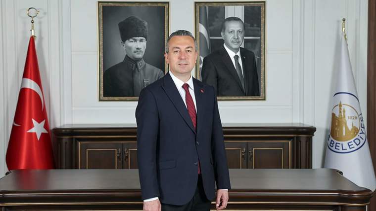 Bir masa önünde duran bir kişi, arkasında iki portre ve Türk bayrağı var.
