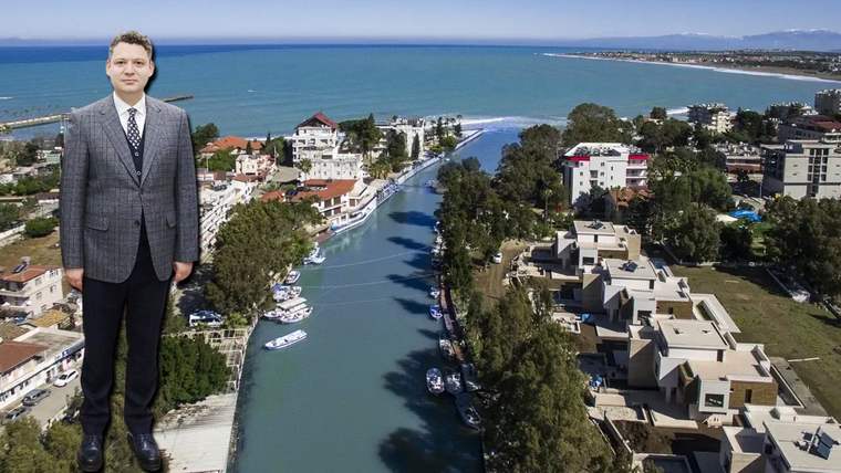 Takım elbiseli bir kişi, teknelerin bulunduğu kanal, çevredeki binalar ve deniz manzarasının önünde duruyor.