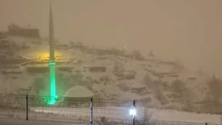 Karla kaplı bir alanın önünde, yeşil ışıkla aydınlatılmış bir minare görünmekte.