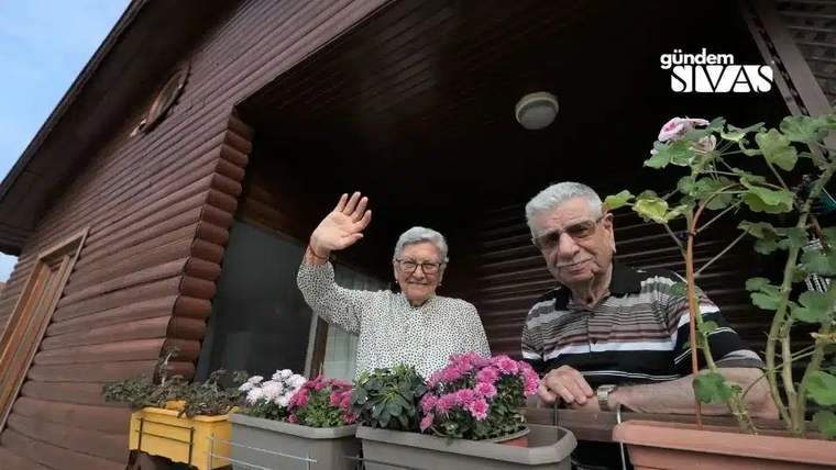 Ahşap evin penceresinden bakan yaşlı çiftin önünde rengârenk çiçek saksıları bulunuyor; kadın el sallıyor.