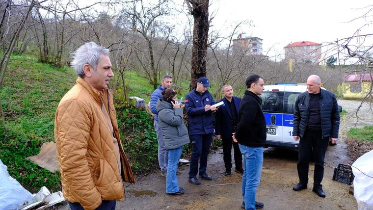 Ağaçlık alanda altı kişi, mavi-beyaz bir araç ve çevredeki kasaların yanında duruyor.
