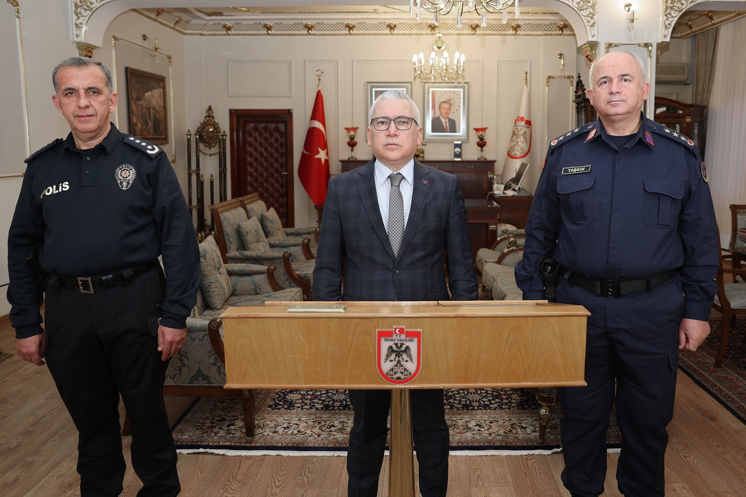 Üç kişi, bir masanın arkasında duruyor; soldaki polis kıyafetli, ortada resmi bir elbise giymiş, sağdaki ise askeri kıyafetli.