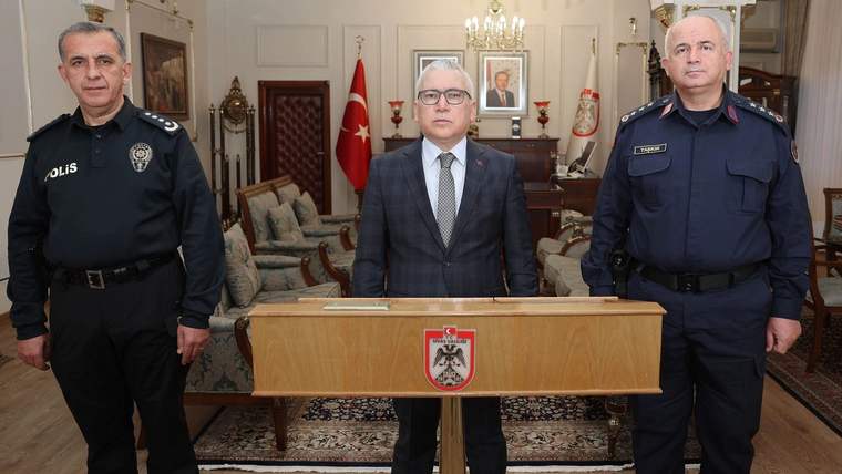 Üç kişi, bir masanın arkasında duruyor; soldaki polis kıyafetli, ortada resmi bir elbise giymiş, sağdaki ise askeri kıyafetli.