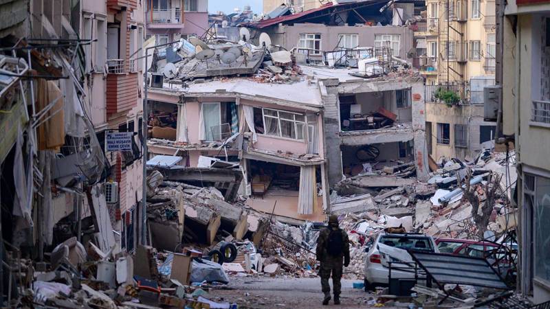 Yıkılmış bir bina ve etrafında dağılmış inşaat malzemeleri, bir kişi yıkıntıların önünde duruyor.