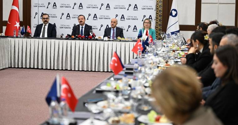 Toplantı salonunda mikrofonlu masada oturan dört kişi, Türk bayrakları ve dinleyicilerle birlikte görülüyor.