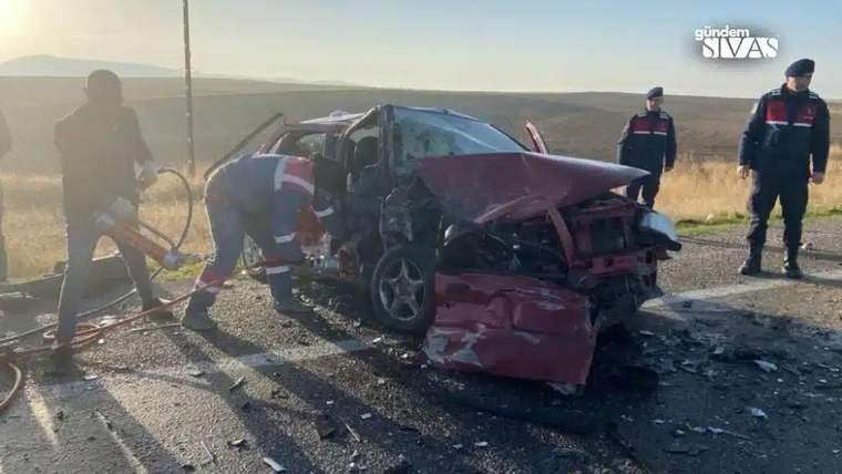 Yol kenarında ağır hasar görmüş kırmızı otomobilin çevresinde üç görevli ve kurtarma ekipmanı görülüyor.