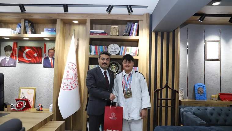 Bir kişi, judocu bir gençle el sıkışıyor; genç, madalyalarını takmış ve judogi giymekte.