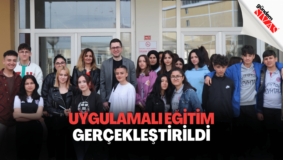 Bir bina girişinin önünde, aralarında iki yetişkinin de bulunduğu kalabalık bir genç grubu birlikte poz veriyor.