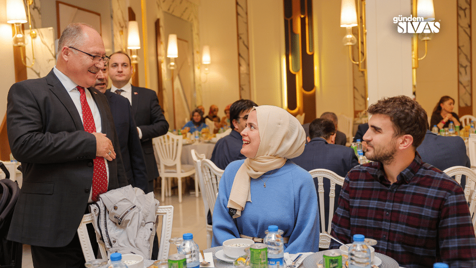 Bir grup insan, yemek masasında oturuyor; bir kişi diğerine konuşuyor.