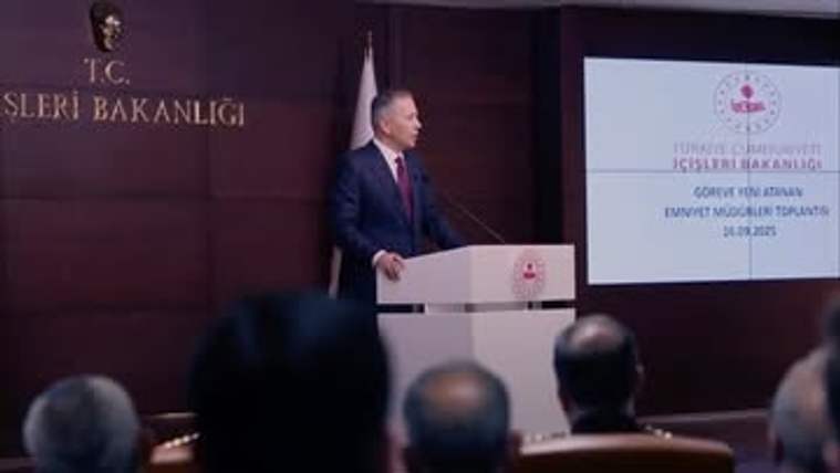 Kürsüde konuşan bir kişi, arkasındaki bayrak ve sunum ekranı önünde salondaki izleyicilere sesleniyor.