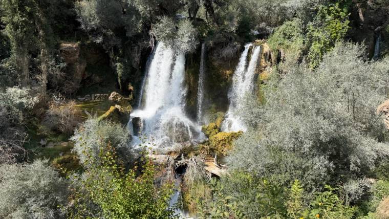 Ağaçlar ve çalılıklarla çevrili kayalık alanda, yan yana akan iki bölümlü şelale görülüyor.