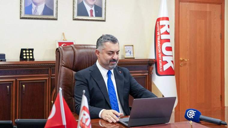 RTÜK bayrakları ve mikrofon bulunan odada bir kişi, masasındaki dizüstü bilgisayarı kullanıyor.