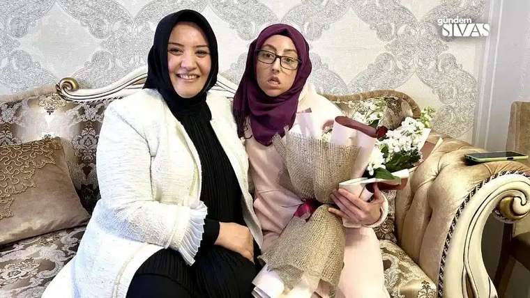 Bir kadın ve engelli genç, bir koltukta gülümseyerek oturuyor; genç, çiçek buketi tutuyor.