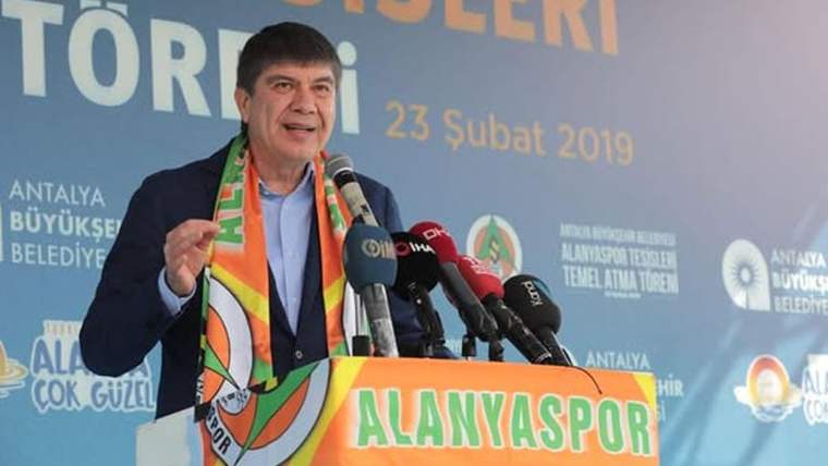 Mikrofonlar önünde konuşan bir kişi, boynunda Alanyaspor atkısıyla elini kaldırıyor; arkada tören afişi bulunuyor.