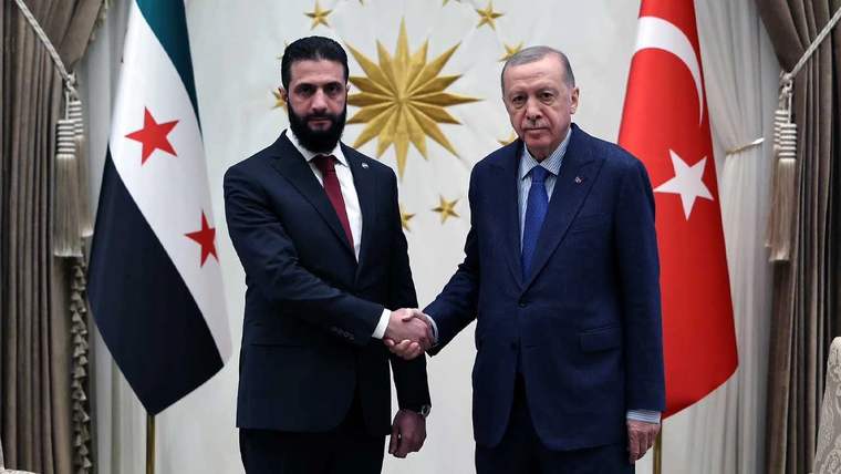 Takım elbiseli iki erkek, Türkiye ve Suriye bayrakları önünde el sıkışırken görülüyor.
