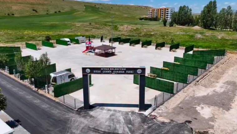 Yeşil çitlerle çevrili alanda çocuk oyun grubu ve banklar, girişte üzerinde yazı bulunan takla yer alıyor.