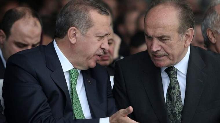 Takım elbiseli iki erkek yan yana duruyor; soldaki el hareketiyle konuşurken sağdaki onu dinliyor.