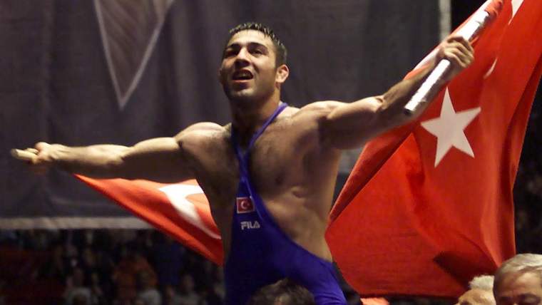 Mavi güreş kıyafeti giymiş bir sporcu, elinde Türk bayrağı tutarak zafer işareti yapıyor.